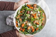 a person holding a white bowl filled with vegetables and grains on top of a table