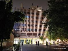 two people walking in front of a tall building at night with lights on the windows