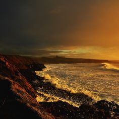 the sun is setting over the ocean with waves crashing on the shore and rocks in the foreground