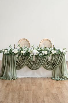 an arrangement of flowers and greenery is displayed on a table with drapes in the middle