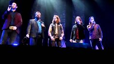 five men standing on stage with microphones up to their ears and one is singing