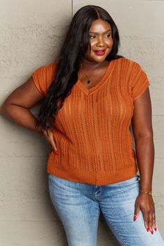 a woman leaning against a wall with her hands on her hips, wearing jeans and an orange sweater