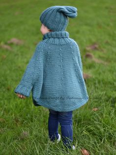 a young child wearing a blue knitted coat standing in the grass with her back turned to the camera