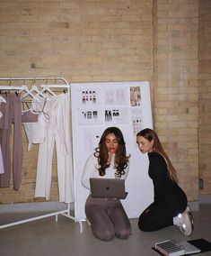 two women sitting on the floor in front of a wall with clothes and laptops