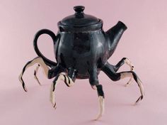 a black teapot with white legs and claws on it's side, sitting in front of a pink background