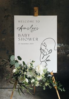 a baby shower sign with flowers and greenery on it next to a wooden easel