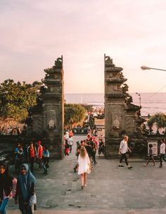 many people are walking around in front of some stone structures near the water at sunset