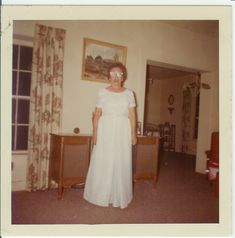 an old photo of a woman in a white dress standing in a living room next to a dresser