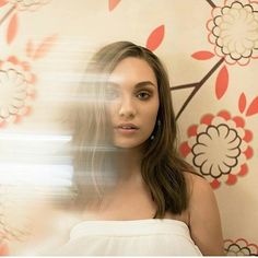 a woman with long hair standing in front of a floral wallpapered background and looking at the camera