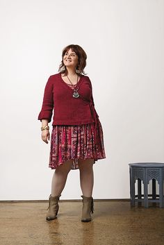 a woman standing in front of a white wall wearing boots and a red sweater over a pink dress