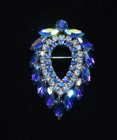 "Bermuda blue, ice blue and blue aurora borealis rhinestones pool in the \"Blue Lagoon\" by D & E for Sarah Coventry, circa 1960s. Large shield shaped brooch in rhodium plate features two inner rows of blue chatons framed by blue ab marquise rhinestones topped with a large blue ab chaton. One of Sarah Coventry's well known, higher end pieces, one can see the influence of DeLizza & Elster's work (famous for it's Juliana line) in the brooch's design, materials, color combinations and workmanship. Formal Blue Cabochon Brooch, Costume Jewelry Blue Brooches With Rhinestones, Formal Blue Gemstone Brooch, Blue Crystal Jewelry Brooch, Blue Crystal Brooch Jewelry, Blue Crystal Jewelry With Brooch Detail, Luxury Antique Blue Brooches, Sarah Coventry Jewelry Vintage Brooch, Blue Crystal Brooches With Rhinestones