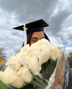 a woman wearing a graduation cap and gown holding flowers in front of her face with cloudy skies behind her