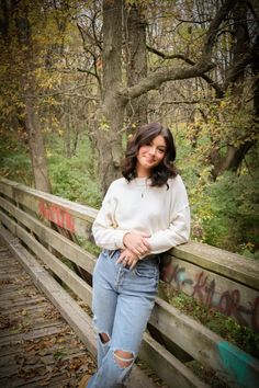 a woman leaning on a wooden fence in the woods with trees and leaves around her