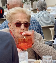 an old woman sitting at a table with a drink in her hand and some food on the table