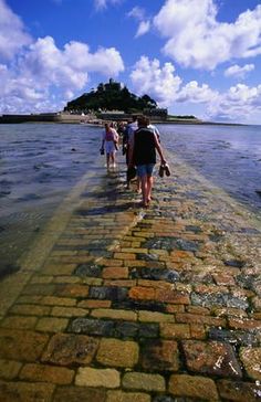 several people walking along the water on a brick path next to an island in the distance