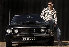 a man sitting on the hood of a black mustang parked in front of a building