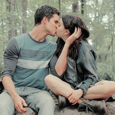 a man and woman sitting on a rock kissing in front of the camera with trees behind them