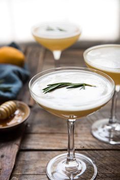 two martinis with rosemary garnish sit on a wooden table next to honey and lemon slices