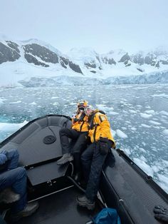 two men in yellow jackets are sitting on a boat near icebergs and mountains