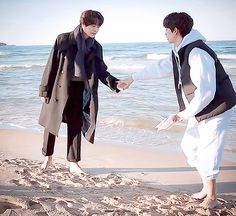 two young men standing on the beach holding hands and looking at each other's hand