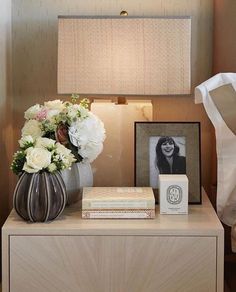 a nightstand with flowers and books on it next to a lamp, photo frame and framed photograph