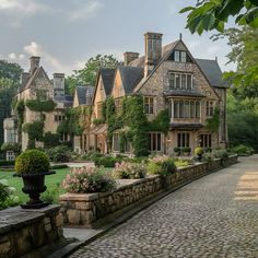 a large stone house surrounded by lush green trees and flowers on a cobblestone walkway