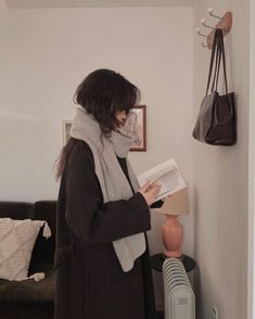 a woman standing in a living room reading a book while wearing a scarf around her neck