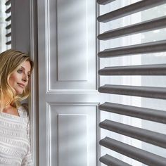 a woman leaning against a wall next to a window with blind shades on the windowsill