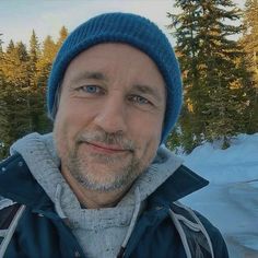 a man wearing a blue hat standing in front of snow covered ground and pine trees