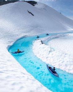 two people are kayaking down a blue stream in the snow with mountains in the background