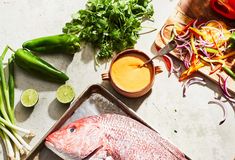 fresh fish and vegetables on a cutting board next to a bowl of sauce, lemons, peppers, onions, celery