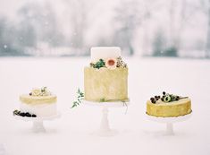 three cakes sitting on top of white pedestals in the snow with trees in the background