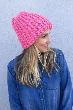 a woman with long hair wearing a pink knitted hat and denim shirt standing in front of a gray wall