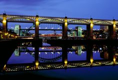 the bridge is lit up at night with lights reflecting in the water and buildings behind it