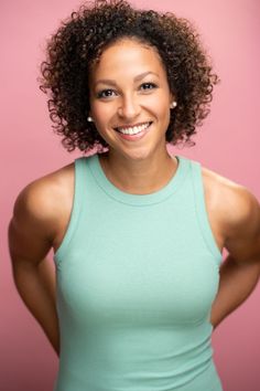 a woman with curly hair wearing a green tank top smiles at the camera while standing in front of a pink background