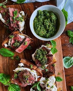 a wooden cutting board topped with slices of meat and veggies next to a bowl of pesto