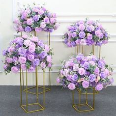 three gold stands with purple flowers on them in front of a white wall and grey carpet