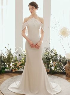 a woman standing in front of a window wearing a wedding dress with an open shoulder