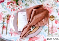 a place setting with pink napkins and goldware on a floral tableclothed table cloth