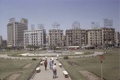 two people walking down a sidewalk in front of some buildings and a park with benches