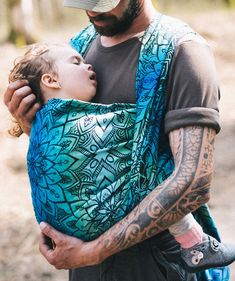 a man holding a baby wearing a blue and green patterned sling on his back in the woods