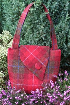 a red plaid purse sitting on top of a purple flower bed in front of a bush