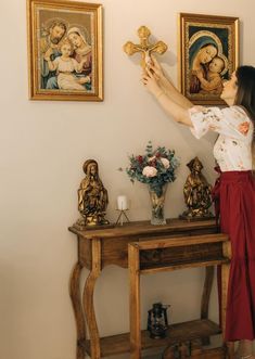 a woman in a white shirt and red skirt holding a cross above a table with paintings on the wall