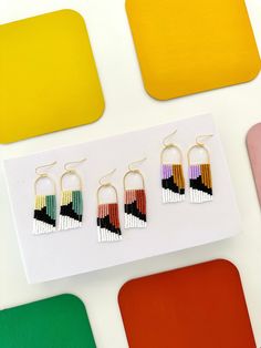 four pairs of earrings with different colors and shapes on white paper next to colored squares