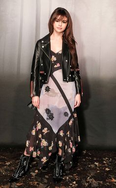 a woman standing in front of a wall wearing a black leather jacket and floral dress