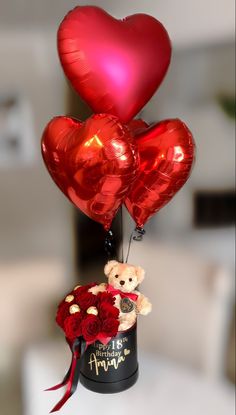 three heart shaped balloons are attached to a hat with a teddy bear and roses in it