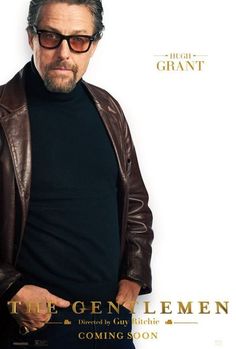 a man in black shirt and brown leather jacket standing next to a white wall with the words the gentlemen on it