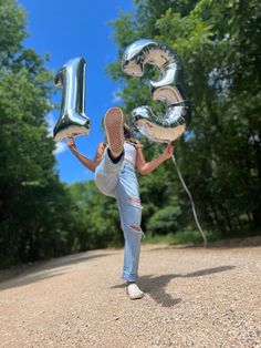 a woman holding up balloons in the air while standing on a dirt road next to trees