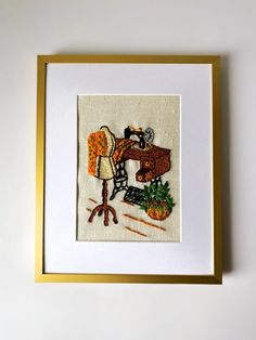 an embroidered picture hanging on the wall with a gold frame and white backing, featuring a sewing machine