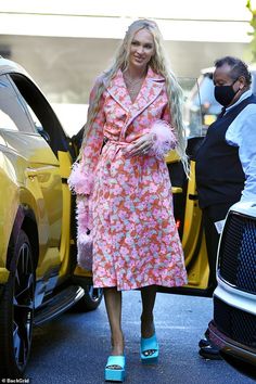 a woman in a pink dress and blue shoes is walking towards a yellow car with her hand on the door handle